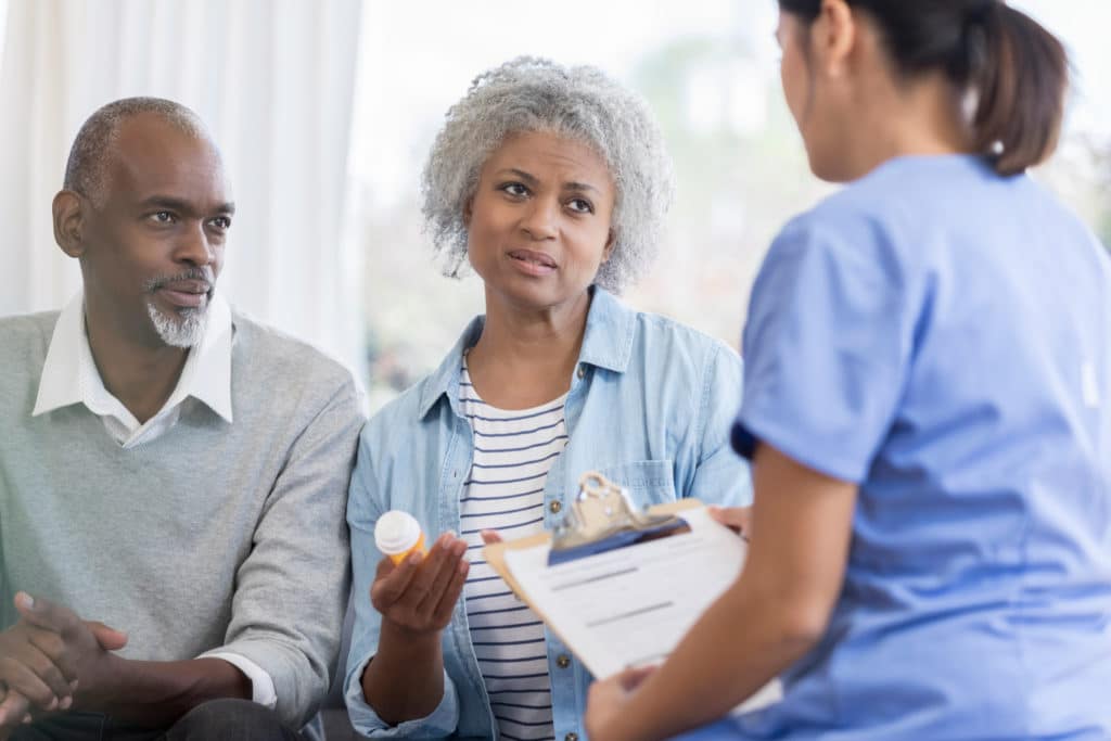 Serious senior African American female patient has concerns about a medication. She holds the medication container while asking a healthcare professional about the medication's side effects.