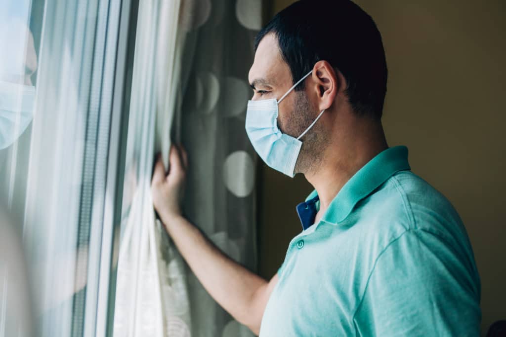 Portrait of a man with protective mask looking through window while he is in home isolation during coronavirus/COVID-19 quarantine.