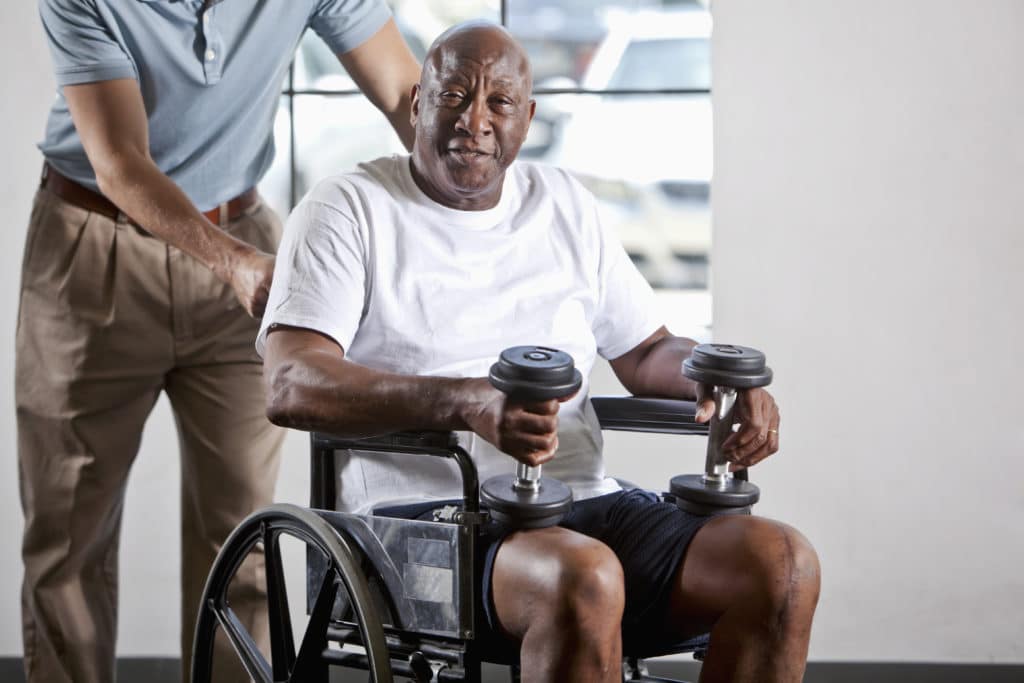 African American man (60s) in wheelchair holding hand weights, ready for physical therapy session.