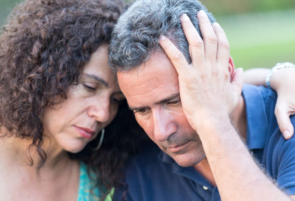 Hispanic mature couple sitting in a park bench, he is very sad and she is consoling him with her arm around him