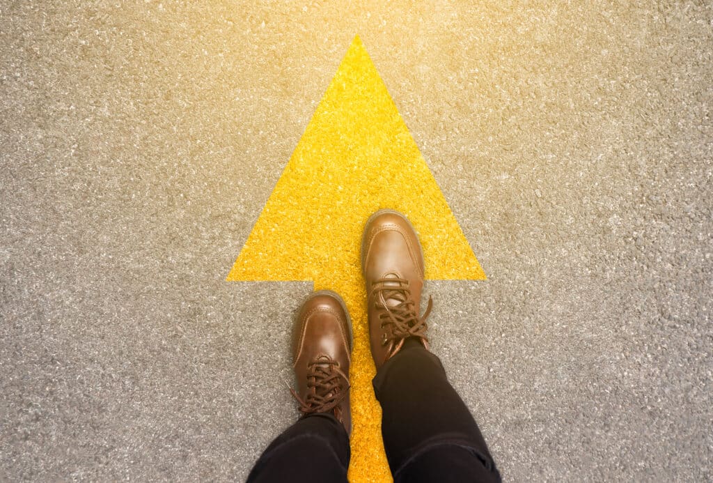 Feet and arrows on road background in starting line beginning idea. Top view. Woman in leather ankle boots on pathway with yellow direction arrow symbol. Moving forward, new start and success concept. Feet and arrows on road background in starting line beginning idea. Top view. Woman in leather ankle boots on pathway with yellow direction arrow symbol. Moving forward, new start and success concept.